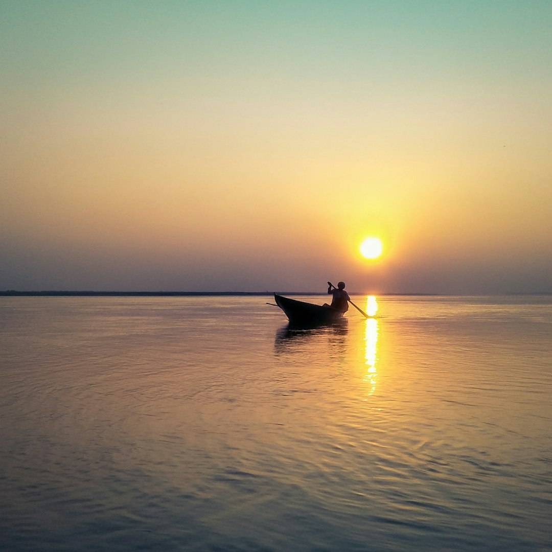 Silhouette of a boatman on the wide Brahmaputra river at golden hour, en route to Majuli island